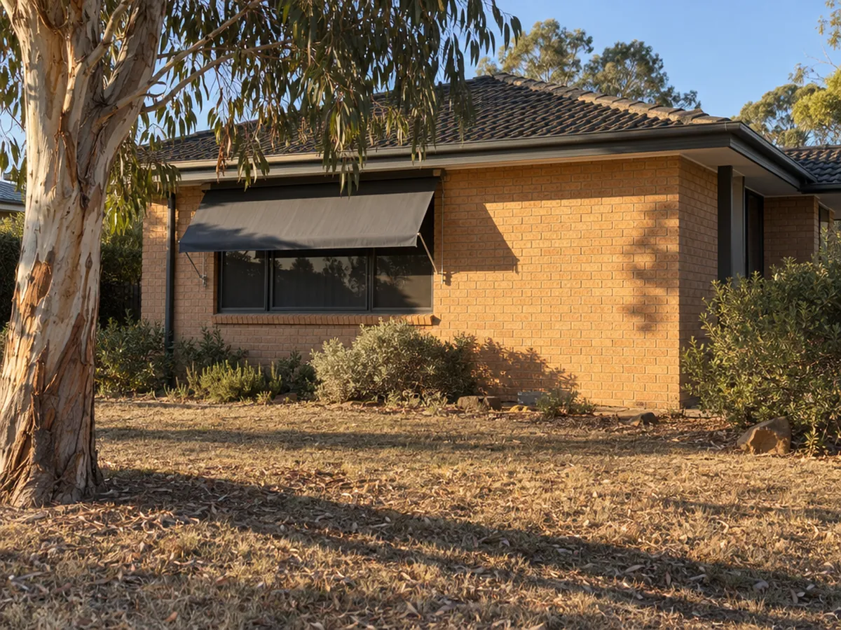 A single-storey brick veneer house in late-afternoon western sun, its fabric awning casting a full shadow over the west window while the bare brick wall beside it sits in bright sun.